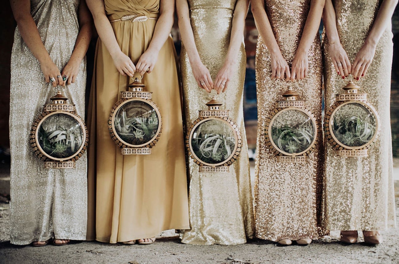 five women in shimmery metallic full-length dresses hold round copper lanterns filled with air plants for a wedding without flowers