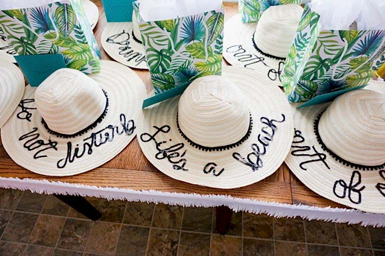 Sun hats with the words "life's a beach" on them, on a table with tropical-themed favor bags for bridal shower favors