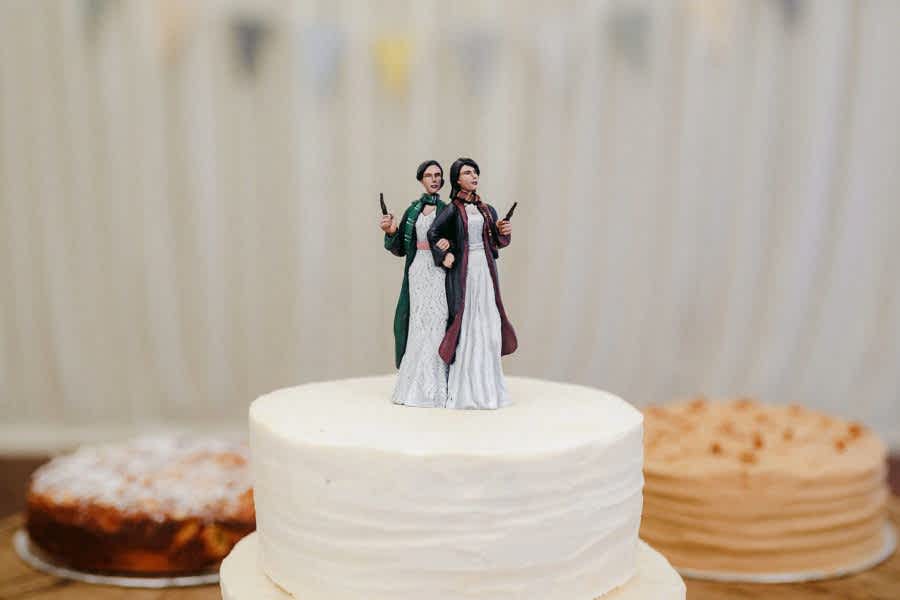 Two womxn in white wedding dresses as cake toppers, one wearing Gryffindor wizarding robes and scarf, the other wearing the same in Slytherin colors. Both hold wands.