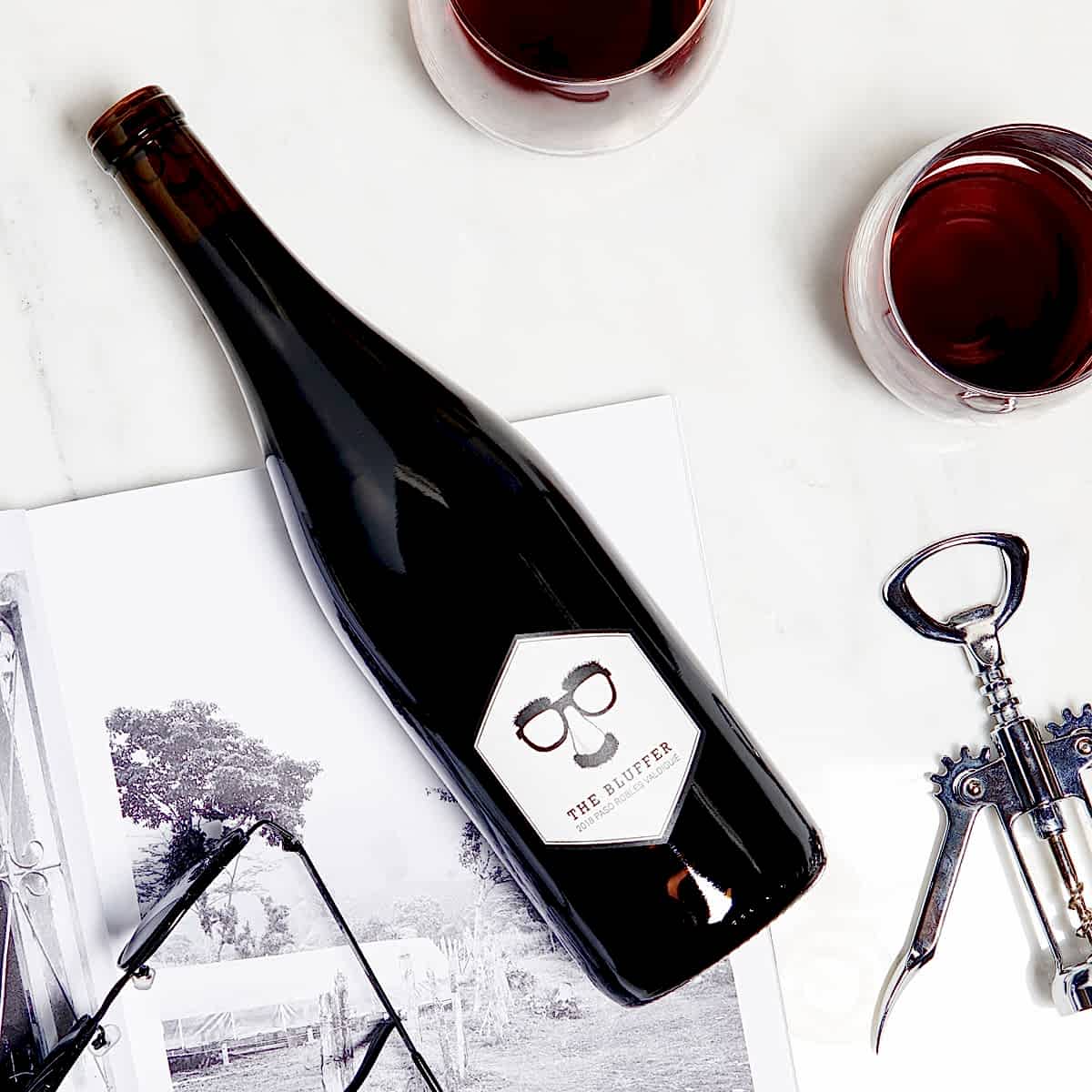 a bottle of Winc's bluffer valdiguié styled in a lay flat on a white surface with a corkscrew, two glasses of red wine, a pencil illustration and a pair of sunglasses