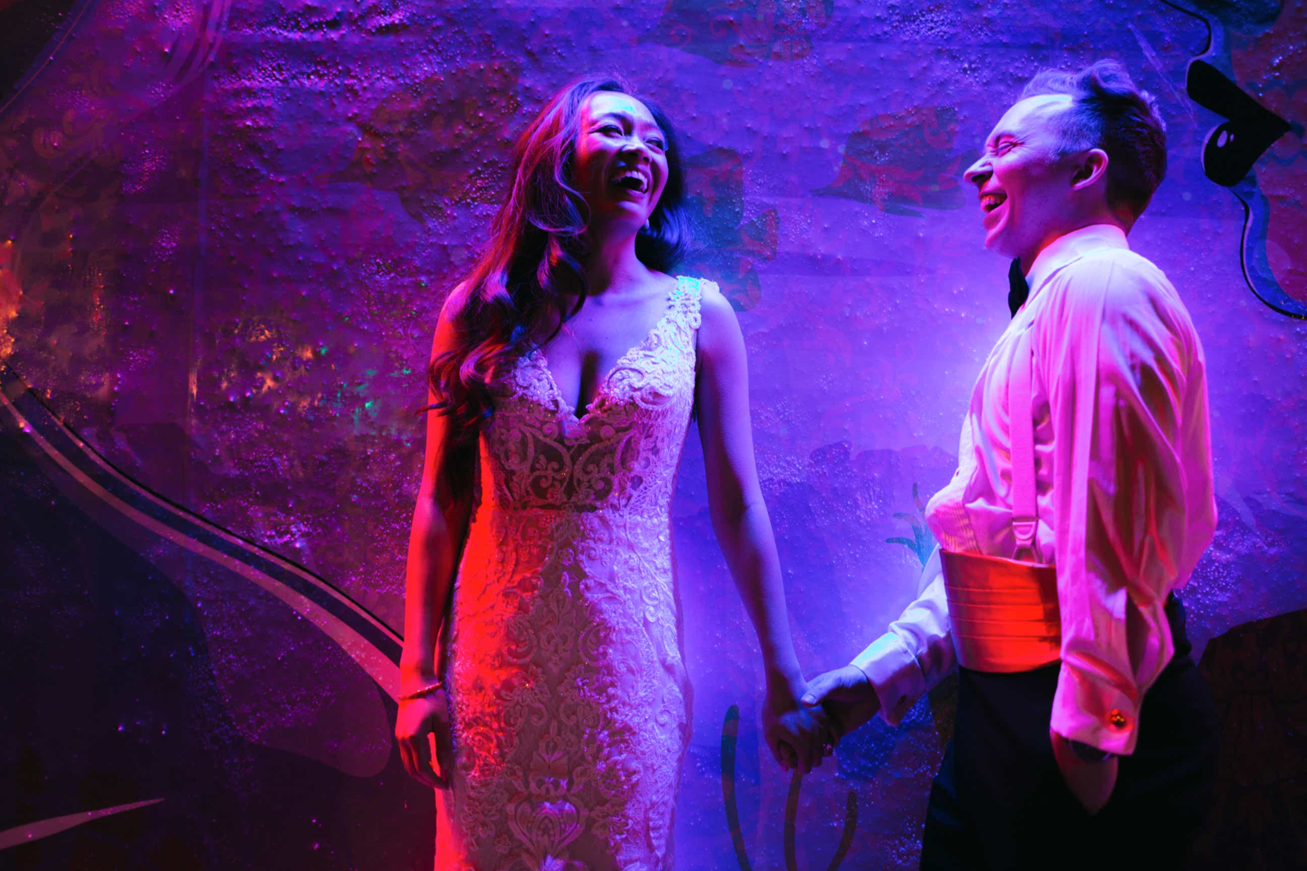 A wedding couple hold hands under purple light.