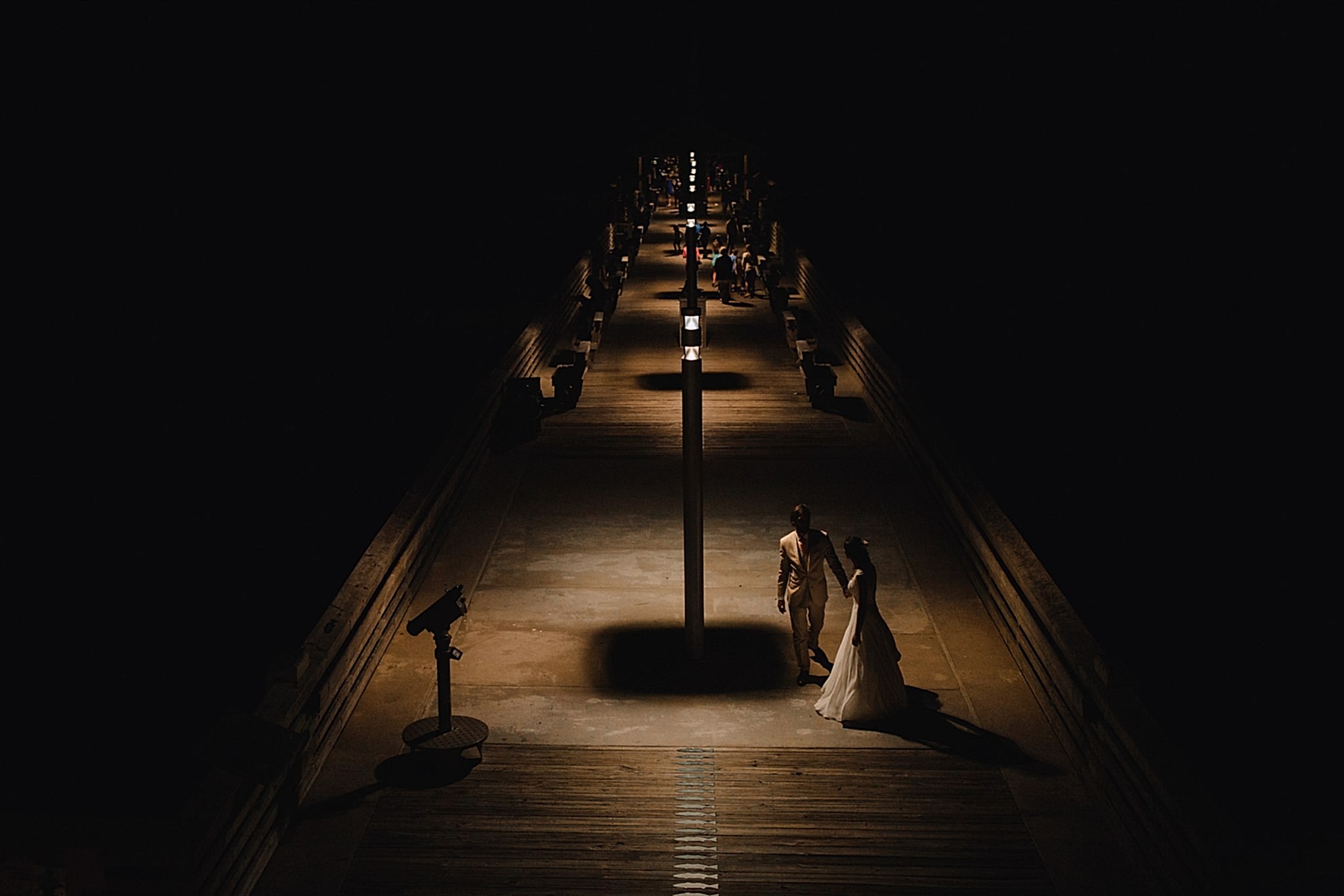 A wedding couple walk along a dimly lit pier at night. Photo by The Commoneer.
