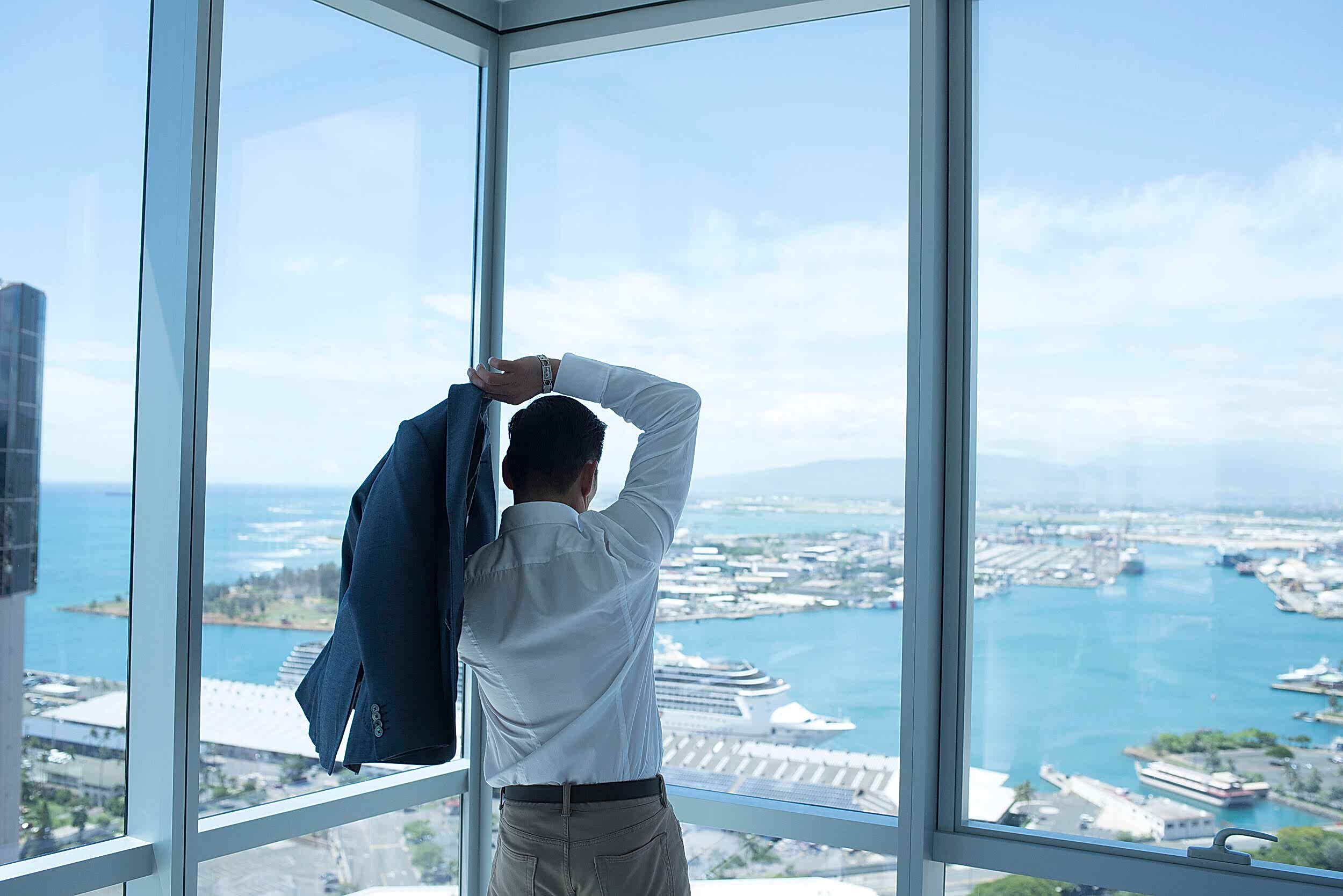 A ma looks out a window and puts on his suit jacket.