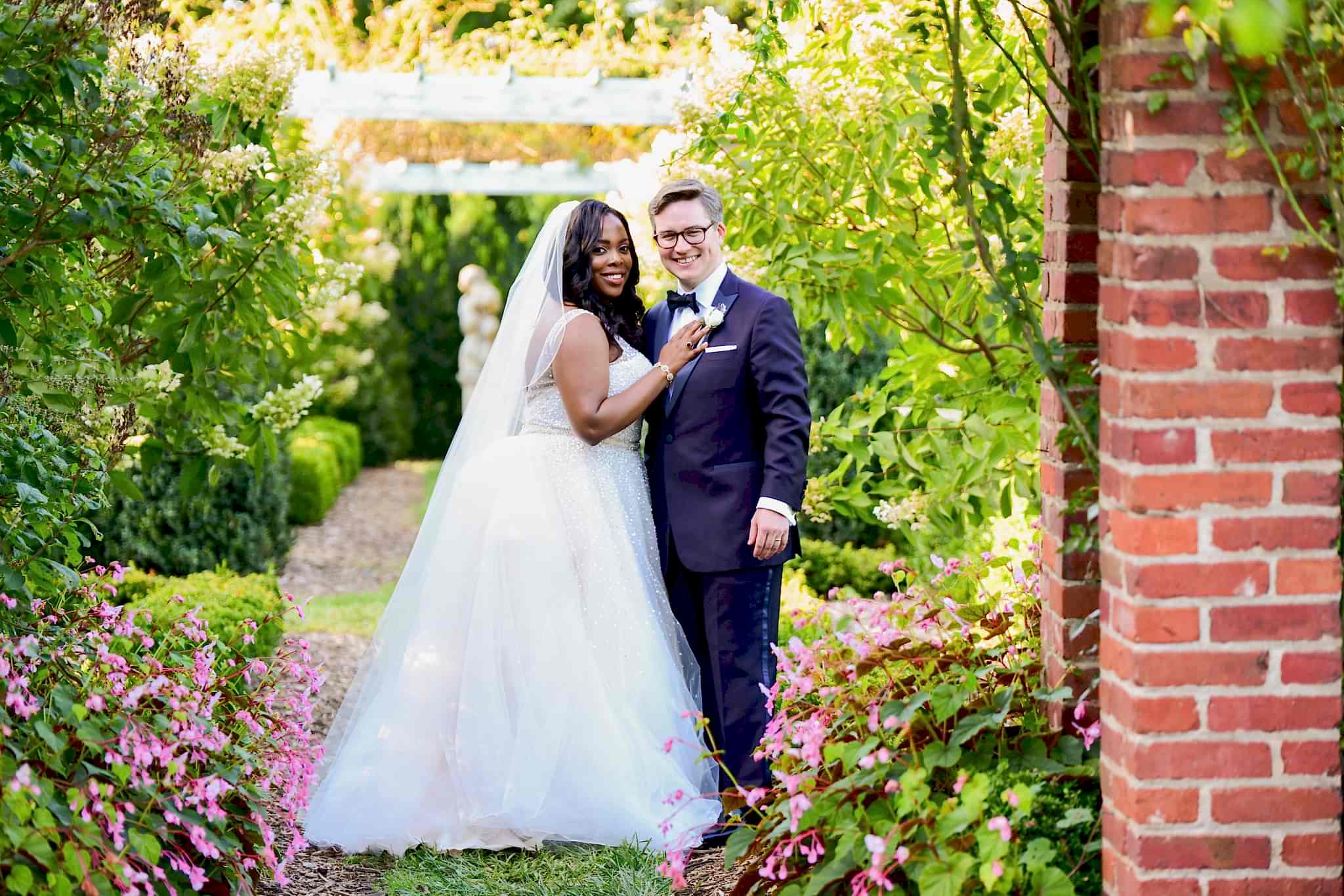 A wedding couple stand and smile on a garden walkway.