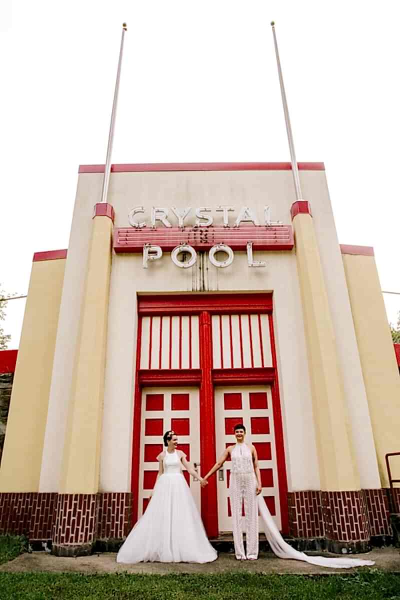 A wedding couple stand outside the gates to a pool and hold hands.