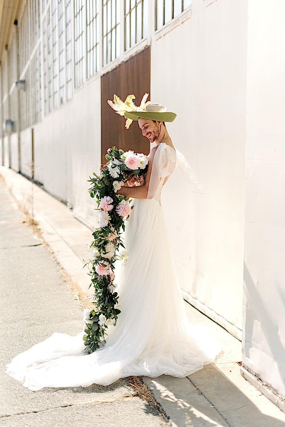 A man wearing a wedding dress smiles at a bouquet.