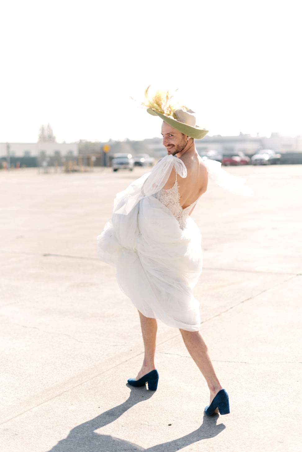 A man wearing a hat and blue heels holds up Maggie Sottero wedding dress style 'Greta' as he walks.