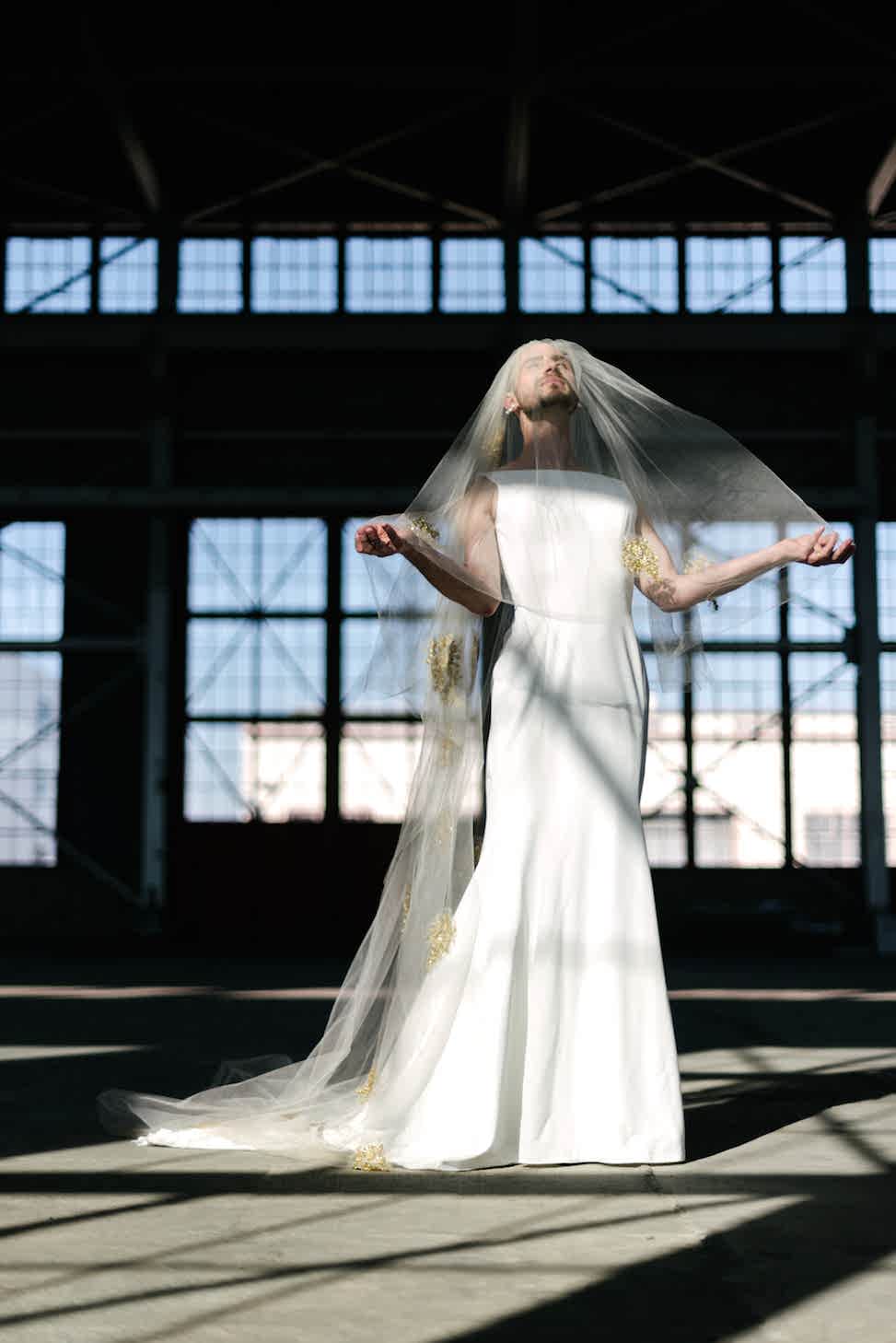 A man stands in a warehouse wearing a veil and Maggie Sottero dress style 'Alice.'