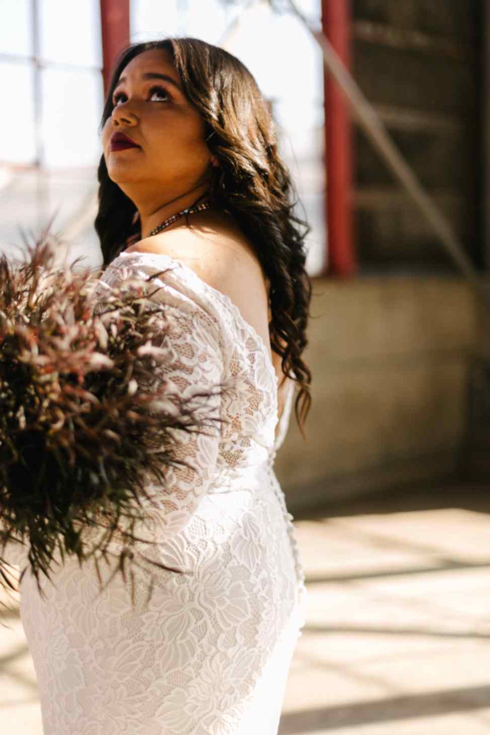 A close-up of a woman with long dark hair wearing a lace wedding dress and holding a bouquet.