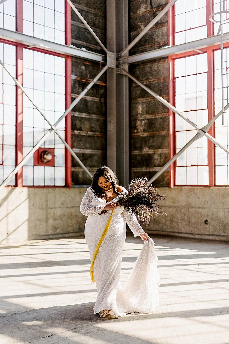 A smiling woman in a warehouse holds a bouquet and wears Maggie Sottero's wedding dress style 'Tina Dawn.'