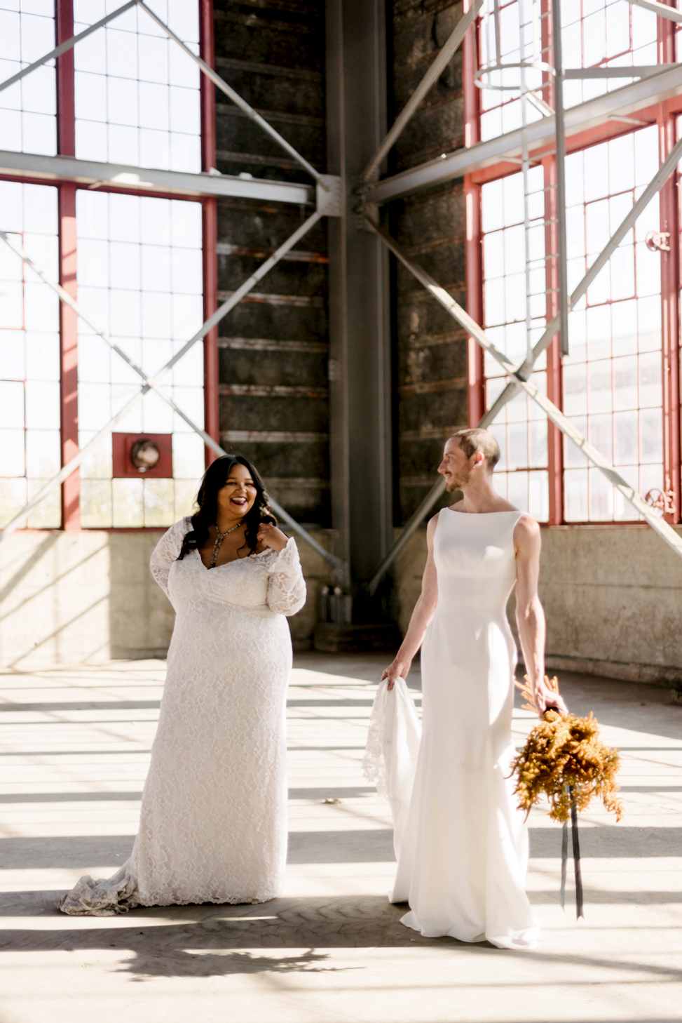 A woman and a man wearing Maggie Sottero wedding dresses stand inside a warehouse.