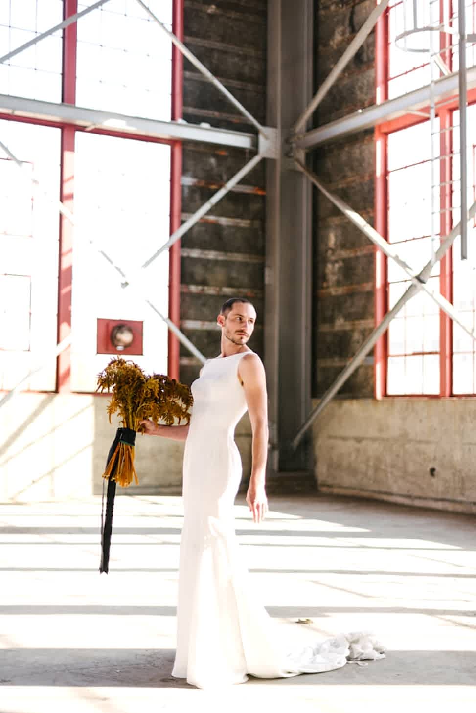 A man standing in a warehouse holds a bouquet and models Maggie Sottero's wedding dress style 'Alice.'