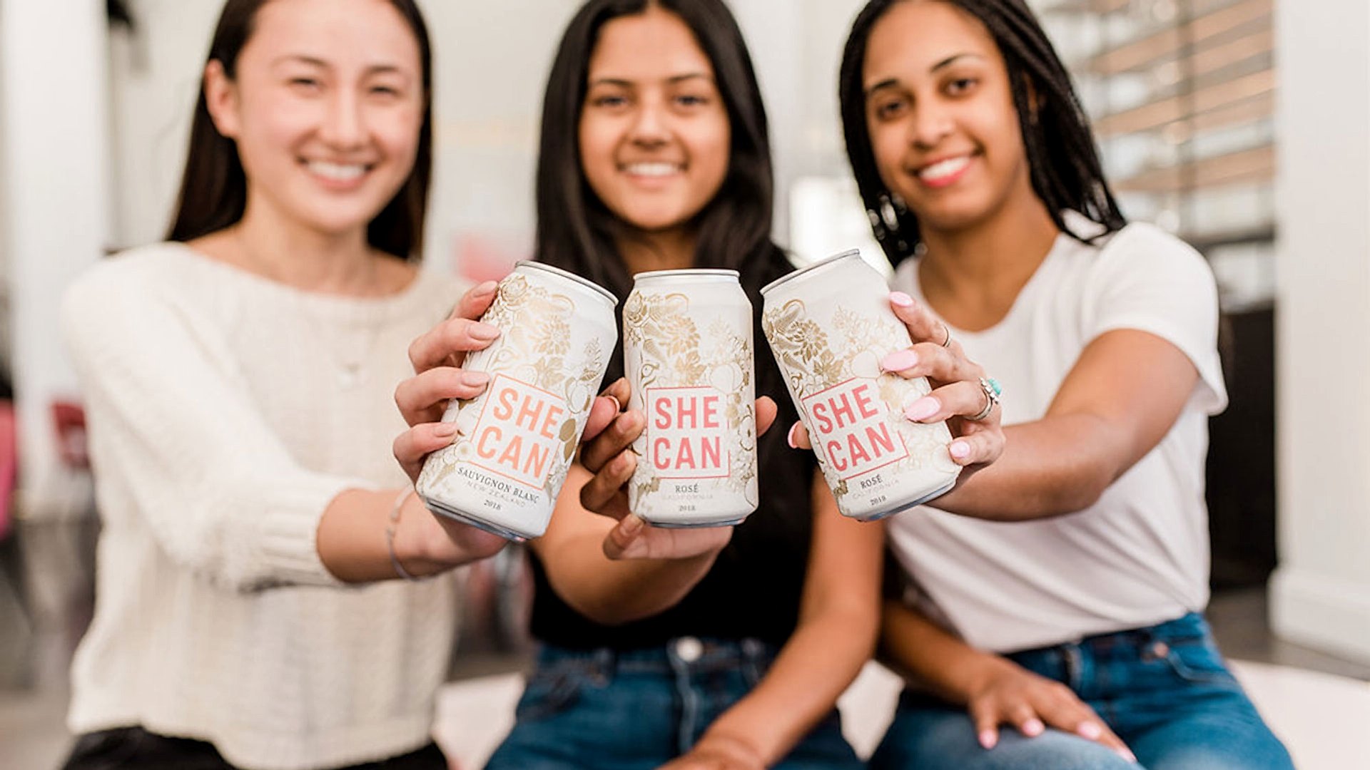 three womxn holding up "SHE CAN" wine cans