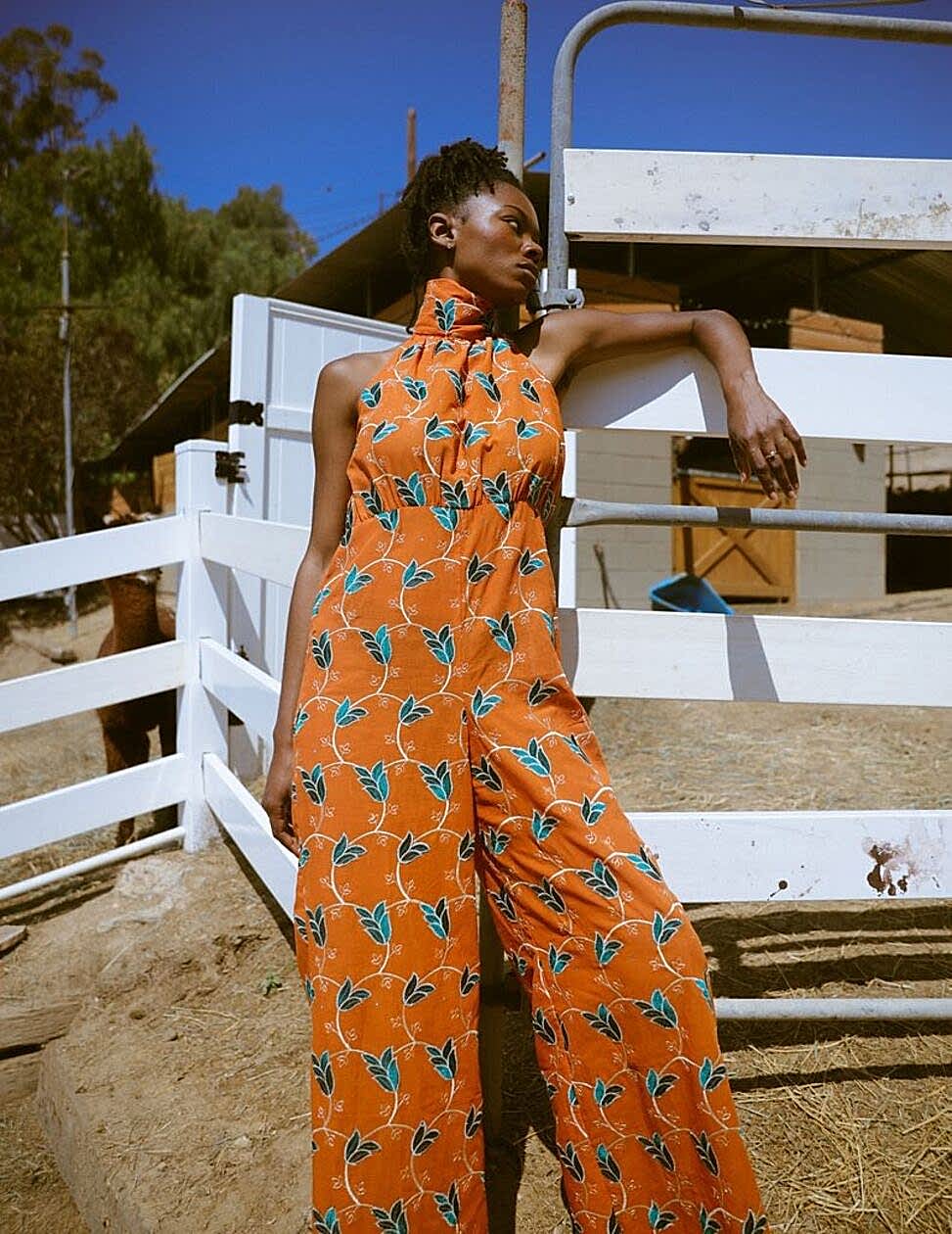 woman leaning against a fence wearing an orange and green leaf printed jumpsuit