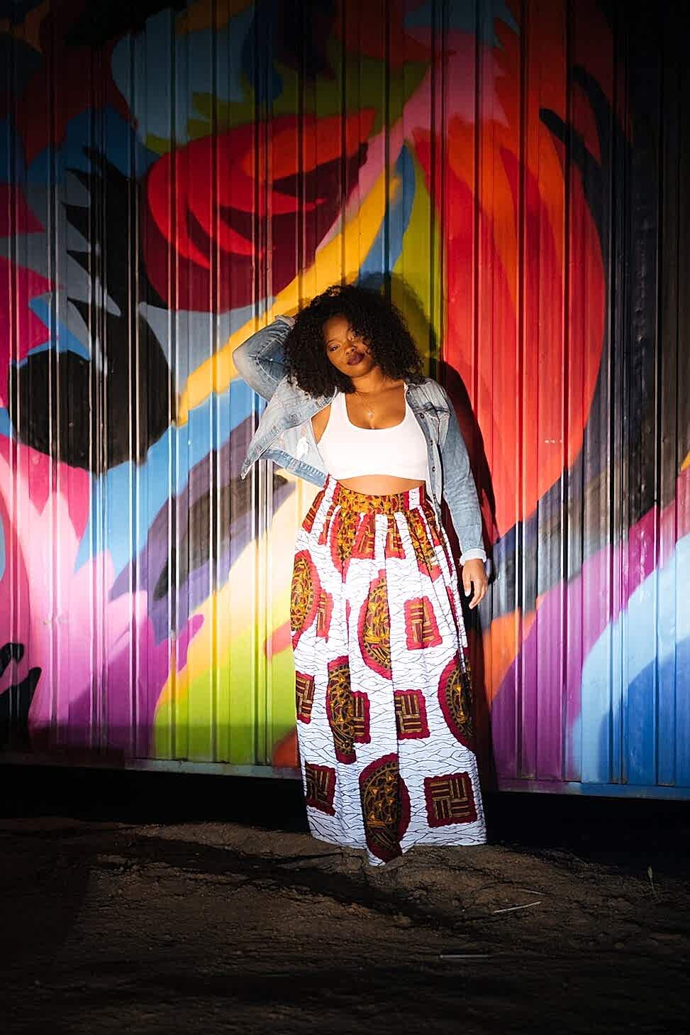 black woman leaning against a graffiti wall wearing a white top with printed pants