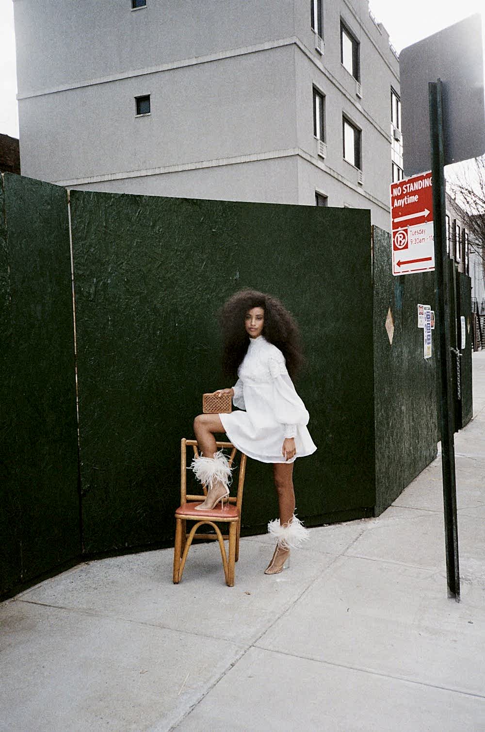 black woman posing in front of a green backdrop wearing a white dress and white feathered shoes
