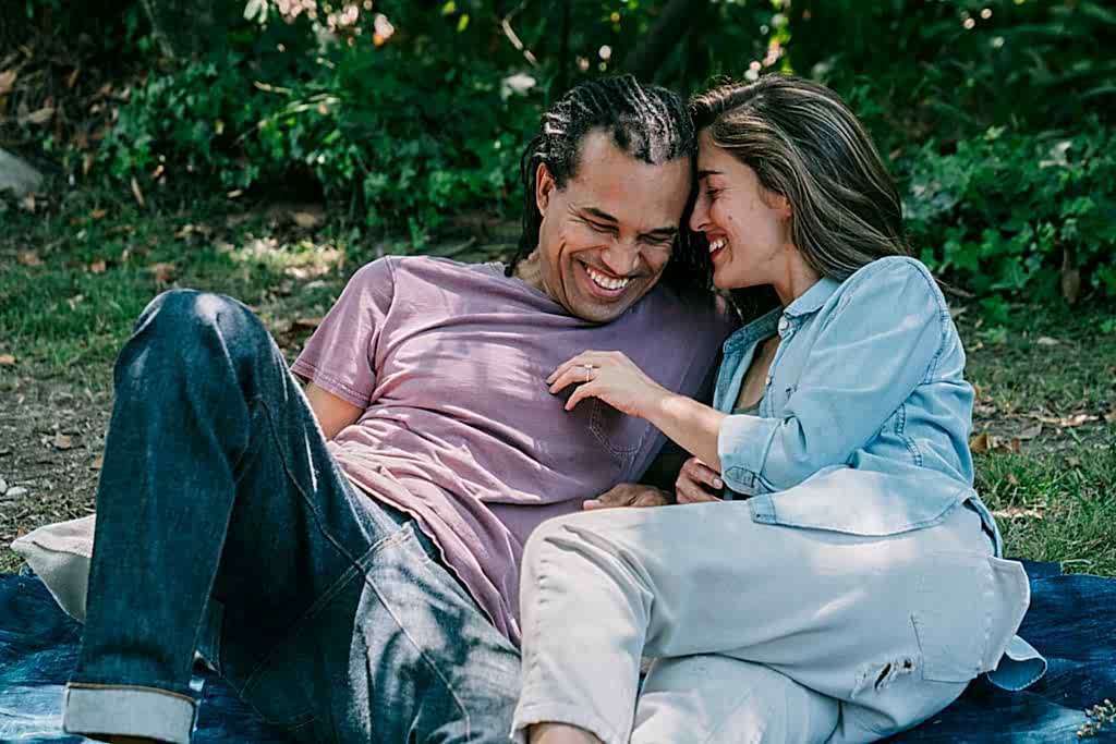 Man and woman lay on picnic blanket, woman's hand with engagement ring shows