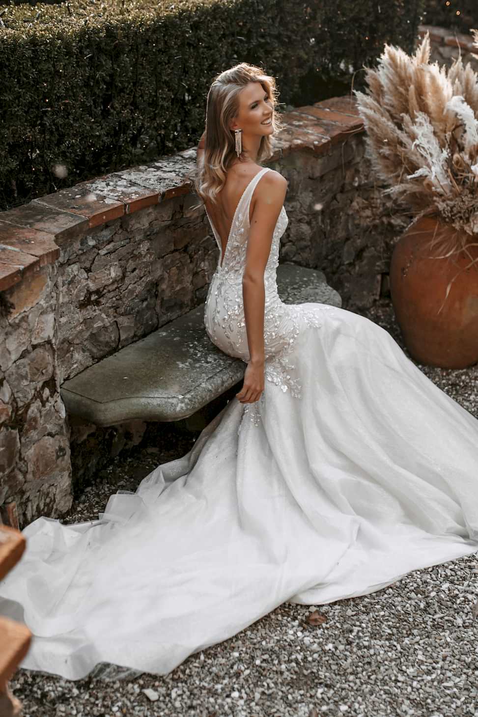 Woman sitting on a bench with low-back, beaded, white wedding dress on, smiling and looking off to the right