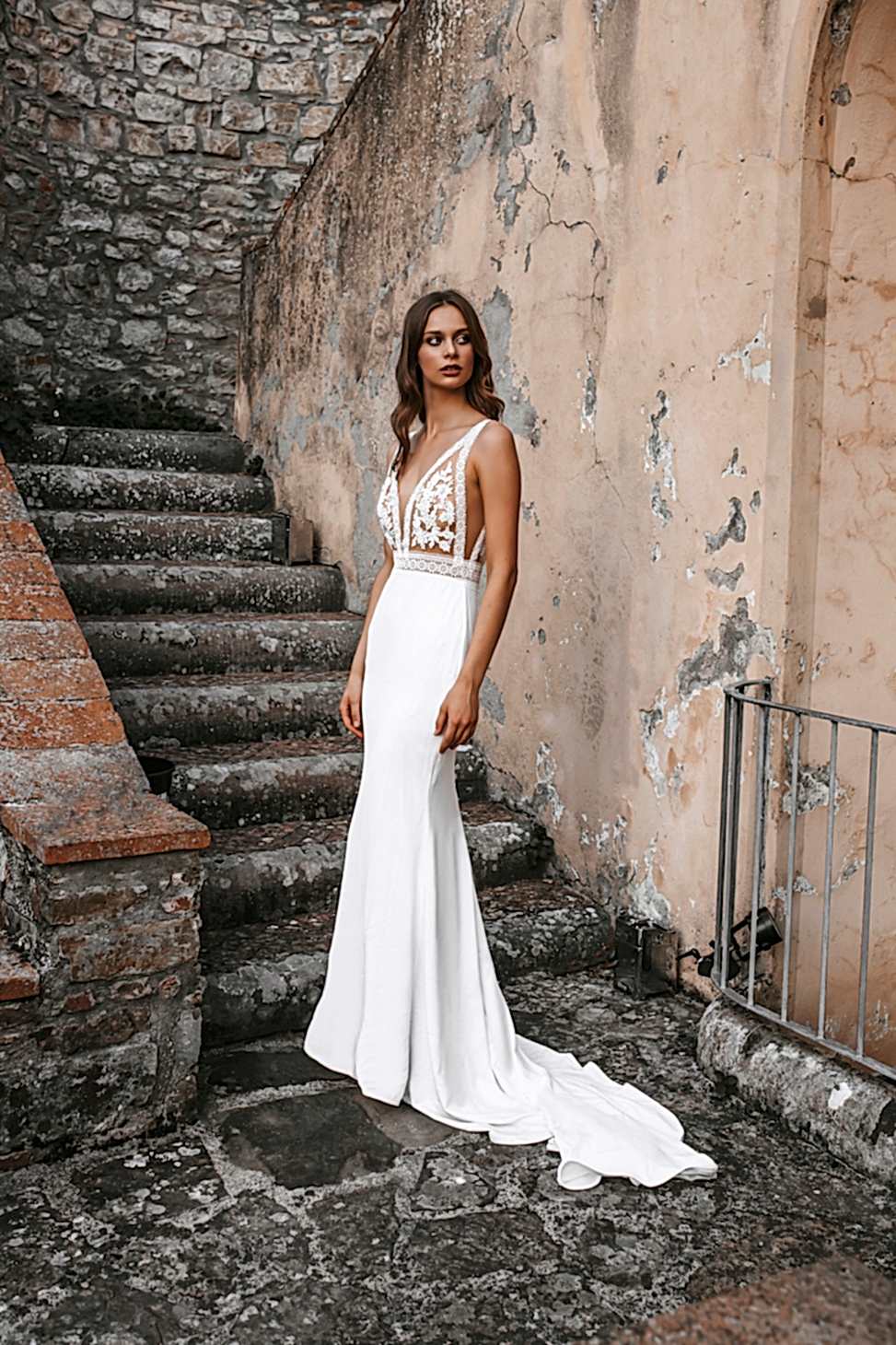 Woman standing in front of run down stone stairs in white wedding dress with sheer and floral top