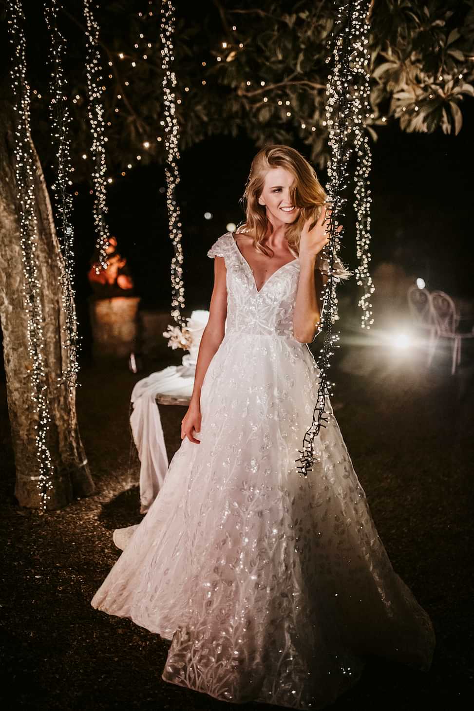 Woman outside among string lights, smiling, wearing a shimmery short sleeved white wedding dress from Allure Bridal