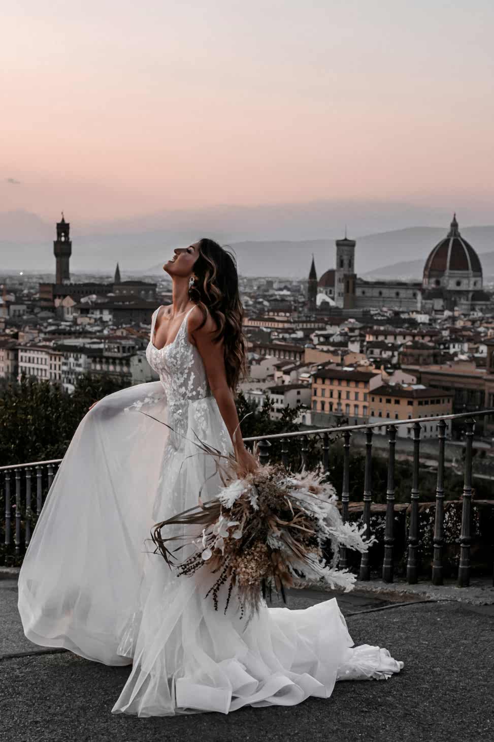 Woman standing in front of city scape, looking to sky, holding up the skirt of white wedding dress in one hand, dried flowers in the other