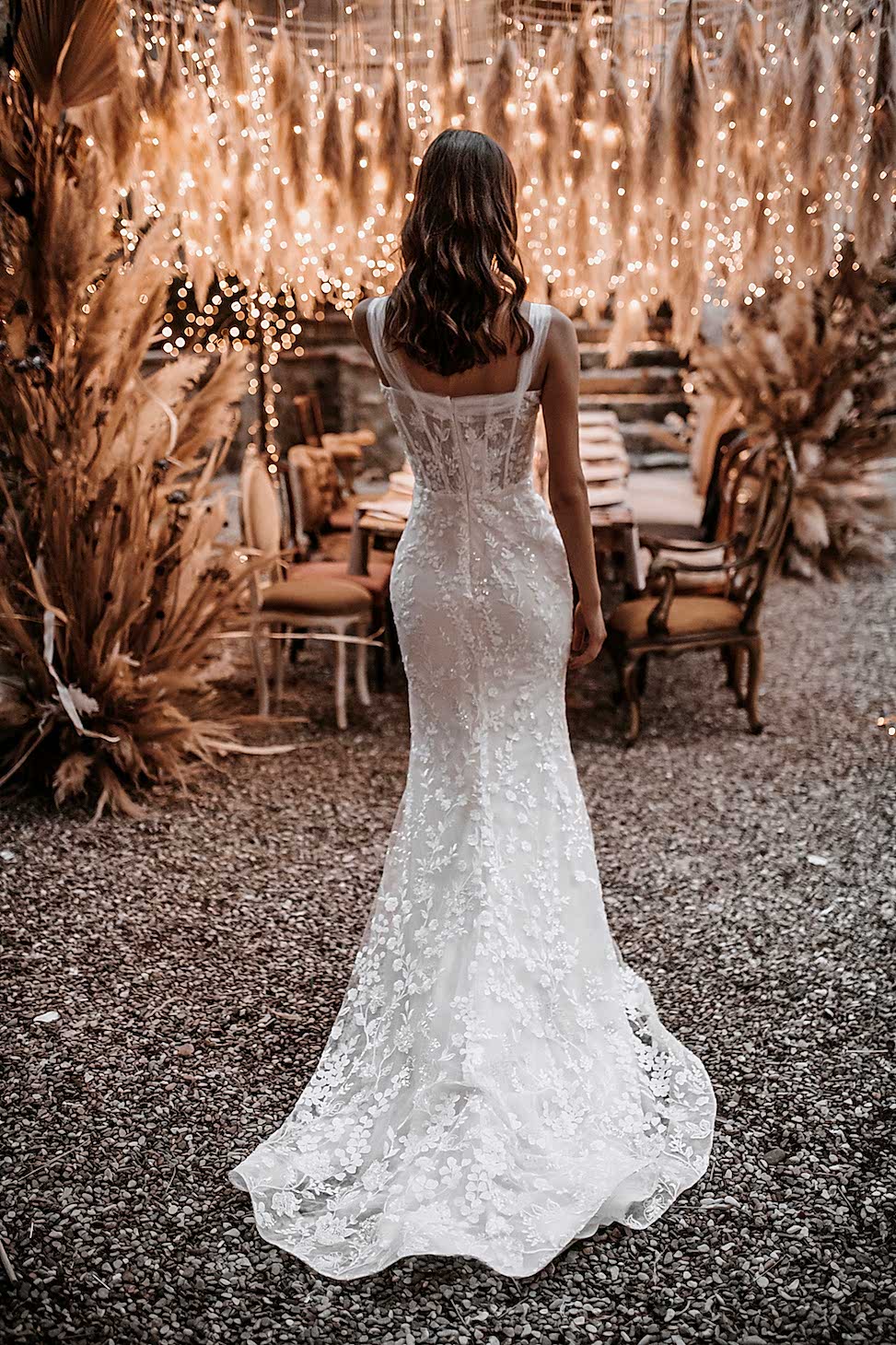 Woman standing in front of outdoor dining table, lookin away, surrounded by dried florals and twinkle lights, wearing a beaded white dress with floral design