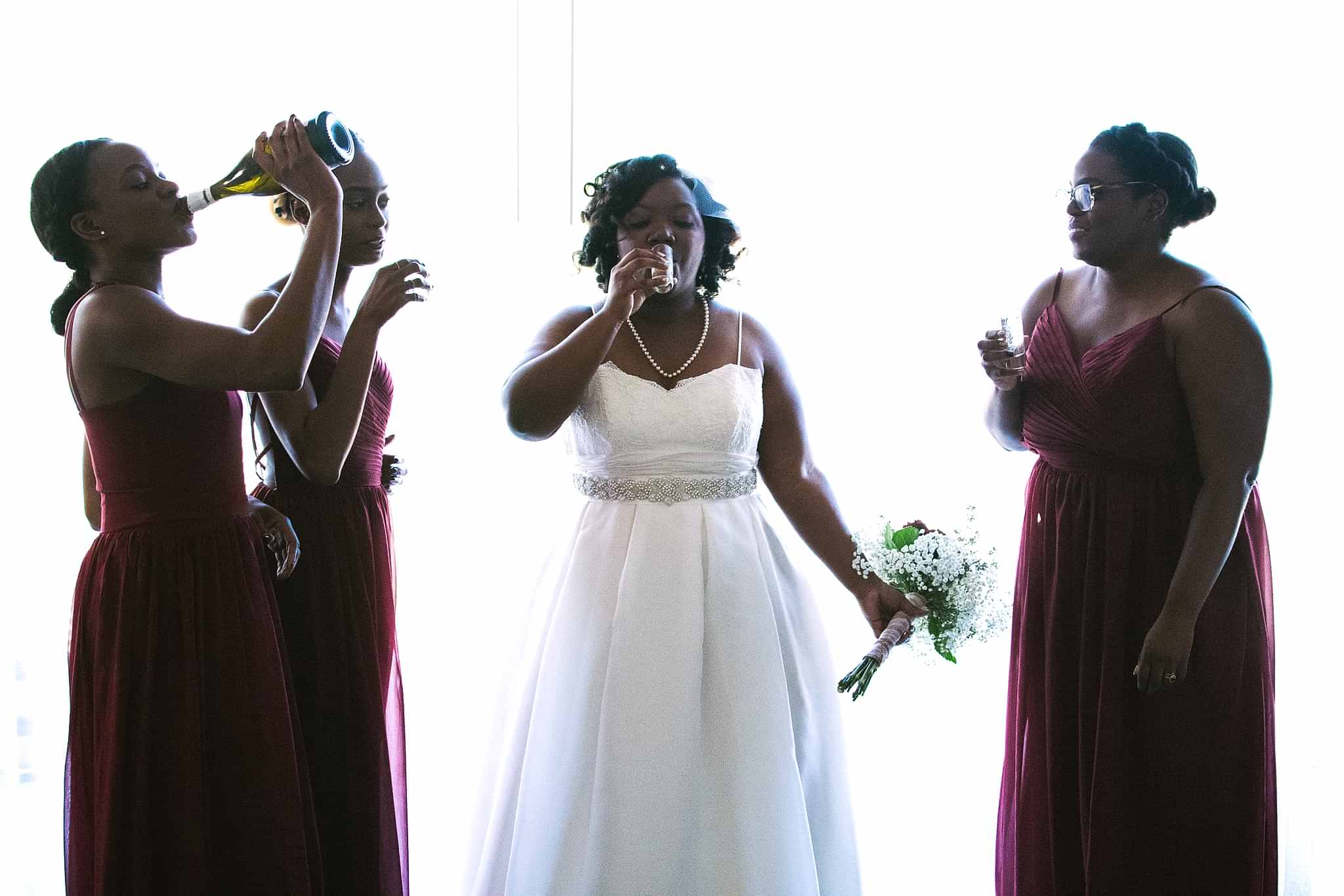 Bride and three bridesmaids stand in front of a window with drinks in hand